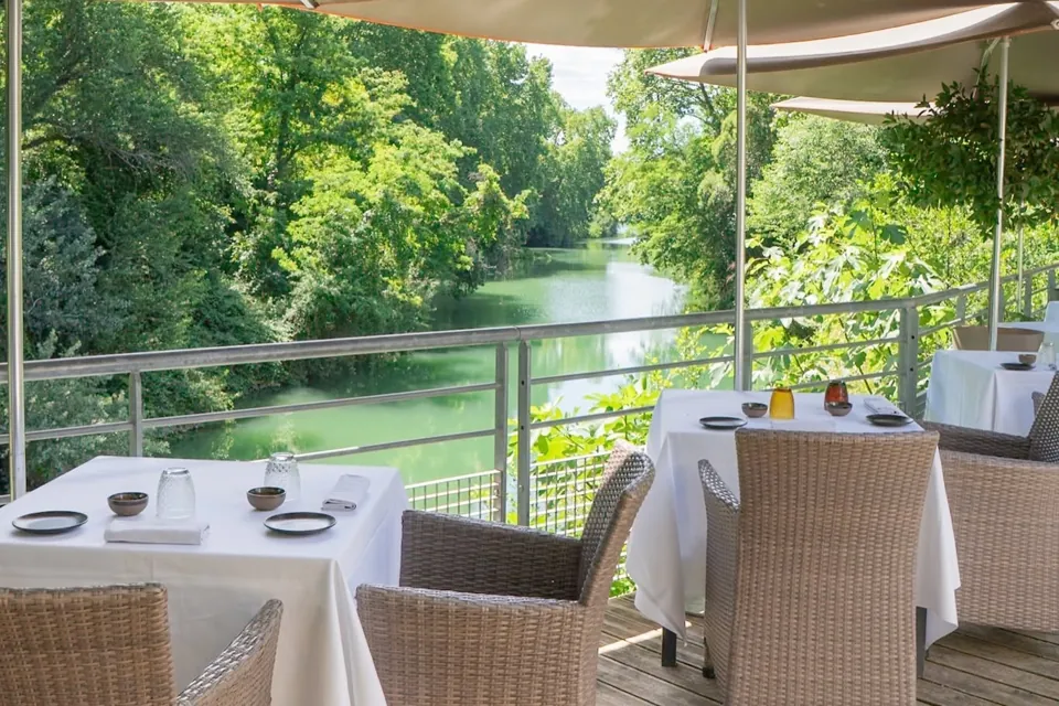 La terrasse en bois sous les parasols, tables nappées de blanc, au-dessus du Lez bordé d'arbres